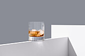 Transparent glass of whiskey on rocks with ice cubes placed on edge of white wooden table isolated on gray background