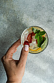 Top view of a cropped hand touching a summer cocktail glass with ice, mint, lemon and strawberry against a grey and white background