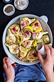 Woman drizzling quartered artichokes in a round baking tin with olive oil on a dark background