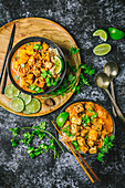 Two bowls of red coconut curry with Tofu and Butternut Squash with rice noodles, in dark bowls on wood chargers, with sliced limes and fresh coriander