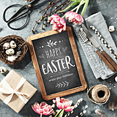 Easter table setting with Happy Easter text on vintage chalk board, spring flowers and cutlery on dark blue background top view flat lay