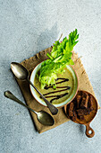 Healthy cream of celery soup in bowl with celery stalks and bowl with slices of bread on napkin with spoons against grey background seen from above