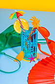 Composition of glass of cocktail and sliced orange fruit with straw, red glasses over surface with inner wear, hand fan on blue and yellow background