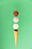 Chocolate, vanilla, strawberry balls and waffle cone on a green background with colourful sprinkles, floating concept. Spring or summer mood