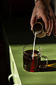 Milk pouring into coffee in glass mug on green table.