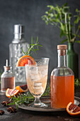 Bright cocktail on ice with blood orange syrup and club soda on ice in vintage glass with syrup bottle, citrus fruits and rosemary garnish