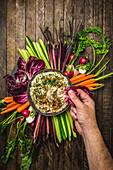 Overhead shot of male hand dipping into onion dip, surrounded by fresh, colorful vegetables on a wood background