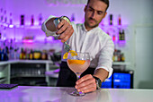 Focused male bartender using tongs to garnish a slice of orange on the rim of a cocktail glass while looking down and standing at the bar in front of blurry blue lights and bottles