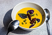 High angle of ceramic bowl with pumpkin cream soup with basil herb, rye bread and seeds against blurred background
