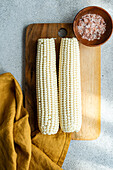 Overhead wooden chopping board with ripe corncobs placed on concrete table in kitchen with bowl with sea salt and napkin