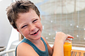 Side view of cheerful cute toothless boy drinking mango juice from glass and looking at camera smiling while sitting at table in cafe during summer vacation