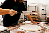 Crop faceless mature male chef in casuals sifting flour with sieve on raw dough over wooden table while making pastry in kitchen at bakery