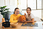 Cheerful young Hispanic female friends looking at camera while sitting at table with Halloween kit bowls and carving eye with sharp tool on orange pumpkin