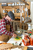 Side view of a mature chef in apron and beret standing on a rustic kitchen focusing intently on slicing leeks, with neatly organized kitchen shelves and hanging utensils in the background