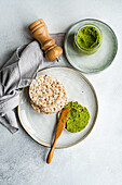 Top view of rice bread on a plate accompanied by a vibrant green spinach pesto pasta-sauce in a glass jar, set against gray backdrop near napkin and salt shaker