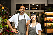 Cheerful young multiracial colleagues in aprons smiling and looking at camera while standing together in floristry store during work process