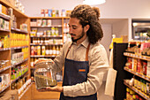 Side view of young ethnic bearded male seller with curly hair in denim apron standing in supermarket with glass jar of bulk food during work