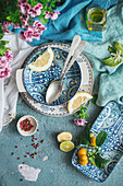 Draufsicht auf einen dekorierten Tisch mit verschiedenem Geschirr und Besteck mit frischen Zitronenscheiben auf blauem Betonhintergrund neben Serviette und Besteck mit Blumenstrauß