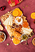 Draufsicht auf eine Gourmet-Käseplatte mit Früchten, Nüssen und Honig, perfekt für die Festtagsbewirtung, auf einem strukturierten roten Hintergrund