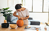 Focused young Hispanic female looking down while standing at table with Halloween tools and pulling out pulp from top of orange pumpkin in daylight