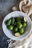 A collection of green feijoa fruits displayed on a white ceramic plate, with a half-cut feijoa showcasing the inner pulp, on a rustic textured background.