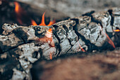 Close-up of glowing embers with small flames, marked by orange hues, in the remnants of a wood fire
