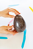 Ausgeschnittene, nicht erkennbare Menschenhand umklammert sanft ein großes gesprenkeltes, dekoratives Ei auf einer weißen Oberfläche mit lebhaften Pinselstrichen