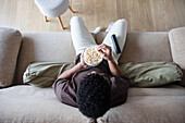 Draufsicht auf einen anonymen, lockig behaarten Afroamerikaner in Freizeitkleidung, der sich auf einem bequemen Sofa mit Fernbedienung ausruht und leckeres Popcorn isst, während er am Wochenende Fernsehkanäle schaut