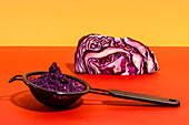 Close up of simple black strainer with red cabbage pulp and half of vegetable for making raw homemade juice on bright background