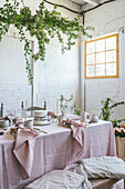 Elegant table served with plates and flowers near yummy cake on pink tablecloth placed near bench covered with cloth against potted plant and white brick wall