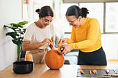 Smiling young Hispanic female friends looking down while standing at table with Halloween tools and pulling out pulp from top of orange pumpkin in daylight