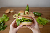 High angle of crop anonymous person holding tasty vegetarian burger with lettuce over blur background with burger burns lettuce and green bottle