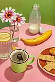 Banana placed near glass bottle of milk and water with lemon slice against flowers in vase and cup of aromatic coffee on pink table against green background