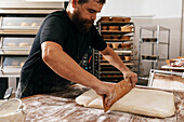 Bearded serious mature male chef with concentration cutting dough with wooden utensil on table while making bread and pastry in kitchen at bakehouse