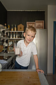 Cute smiling Caucasian boy with blonde hair holding milk glass and looking at camera while cooking food and standing at kitchen counter in house
