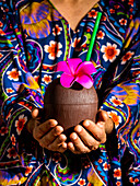 Gesichtsloser Mann in lebhaftem Hemd mit ornamentalem Blumenmuster hält Kokosnusscocktail mit Strohhalm und Blumendekoration auf blauem Hintergrund