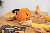 Anonymous women sitting at table and carving with tool scary teeth on orange pumpkin and preparing for Halloween decoration in day