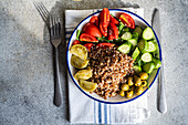 Draufsicht auf eine gesunde Lunch-Schale mit gekochtem Bio-Buchweizen, frischer Gurke und Tomate sowie fermentierter Tomate und Oliven auf einer Serviette zwischen Gabel und Messer auf einer grauen Fläche
