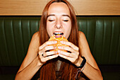 Young woman with red hair and piercing with closed eyes sitting at table and enjoy eating hamburger in restaurant