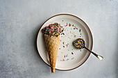 Close up of a waffle cone with coffee and chocolate ice cream with a spoonful of multicolored sugar placed on a ceramic plate on a concrete background