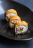 Focused row of tasty fried sushi rolls with cooked rice and salmon with avocado slices on ceramic black plate on table