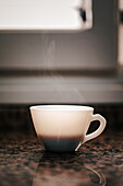 A steaming white coffee cup sits elegantly on a granite countertop illuminated by soft ambient light from above exuding a sense of calm morning ritual