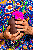 Gesichtsloser Mann in lebhaftem Hemd mit ornamentalem Blumenmuster hält Kokosnusscocktail mit Strohhalm und Blumendekoration auf blauem Hintergrund
