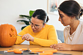 Focused women looking down while sitting at table and carving with tool scary teeth on orange pumpkin and preparing for Halloween decoration in day