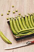 Reife grüne Erbsen in einem braunen rustikalen Tablett neben einem Taschenmesser auf einer hölzernen Tischplatte, mit verstreuten Erbsen drum herum