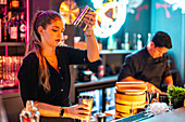 Side view of Beautiful young barmaid with concentration mixing alcoholic drink in cocktail shaker at counter while working in illuminated night pub