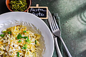 A plate of appetizing pesto Farfalle pasta sprinkled with cheese and herbs, with a fork on the side, and a chalkboard sign.