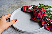 Unrecognizable female putting ceramic plate with fork and knife wrapped in checkered handkerchief on gray table near green fir sprigs