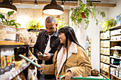 Happy young multiracial couple in casual wear smiling and choosing products while shopping together in modern store with cart