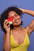 Friendly charismatic African American female model smiling with closed eyes while hiding eye behind slice of sweet watermelon